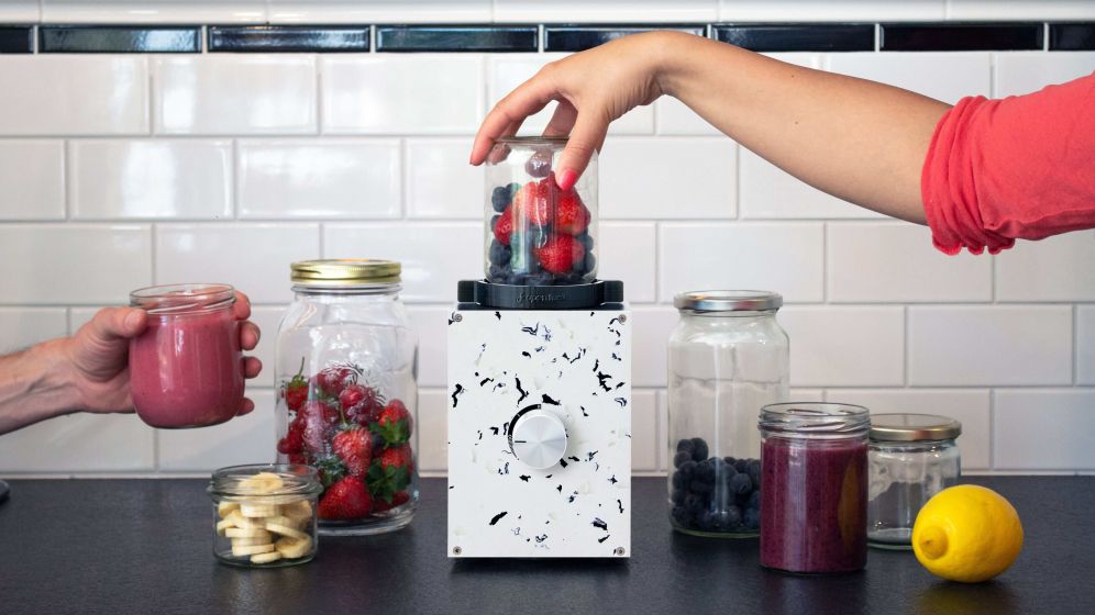 Image of a blender on a kitchen counter filled with fruit, with a hand on the blender. 