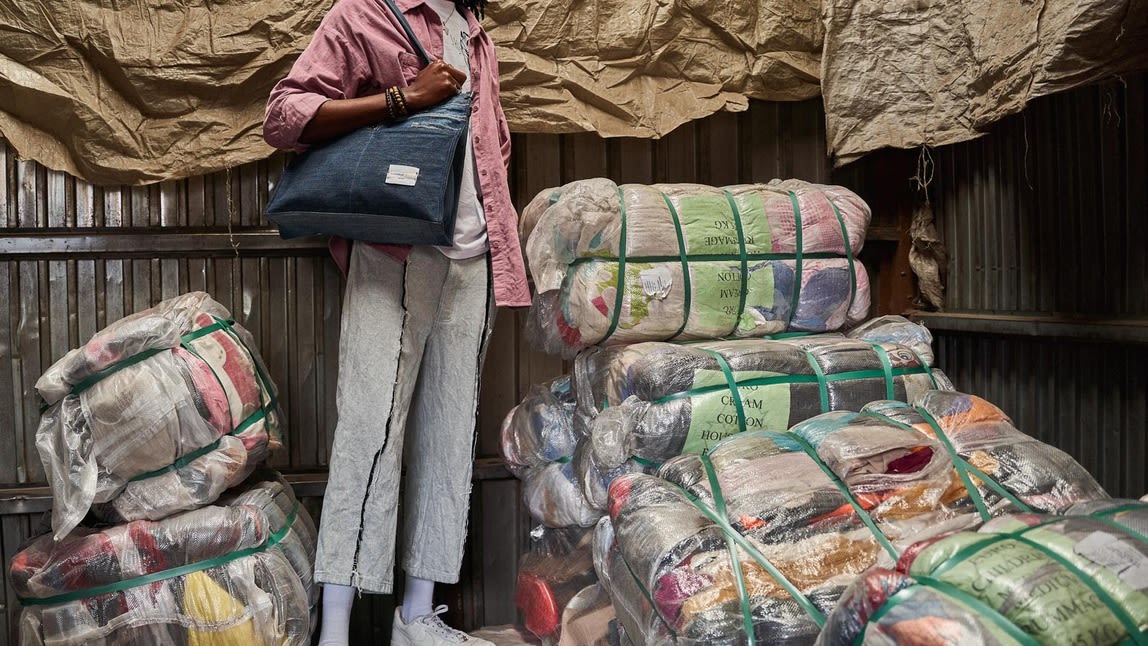 An African man standing with a bag stood next to big packages of reused material